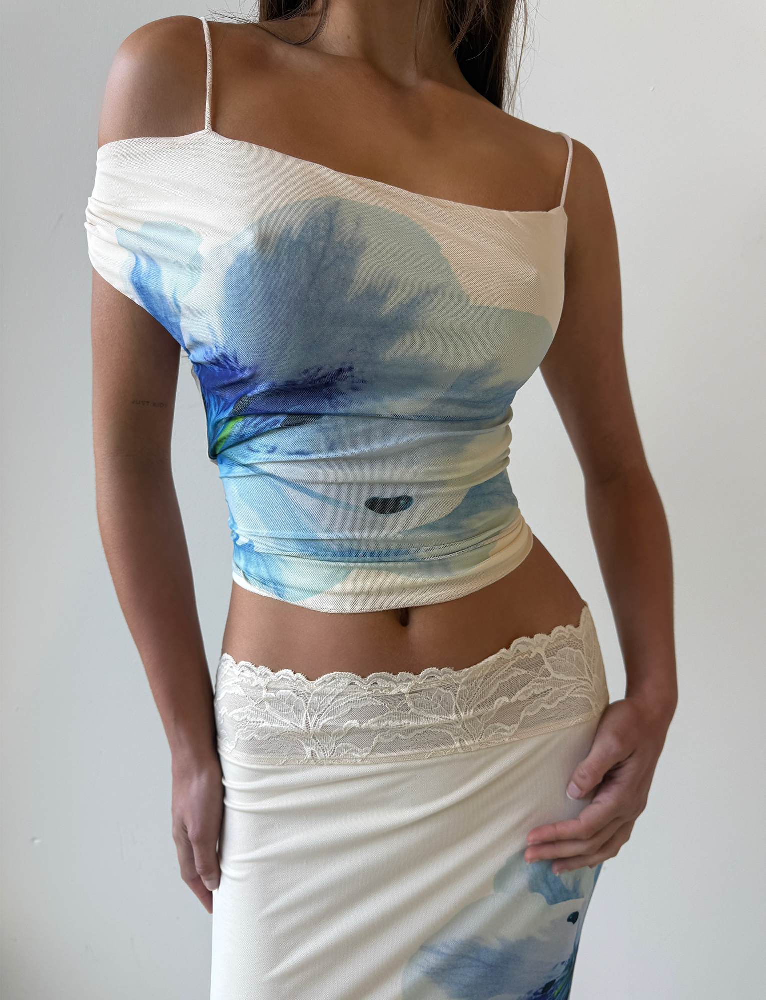A woman models the TIGERMIST LILLIAN TOP in white cream with a floral print, featuring an asymmetrical off-shoulder design and lace waist trim, standing against a plain white background with her face not visible.