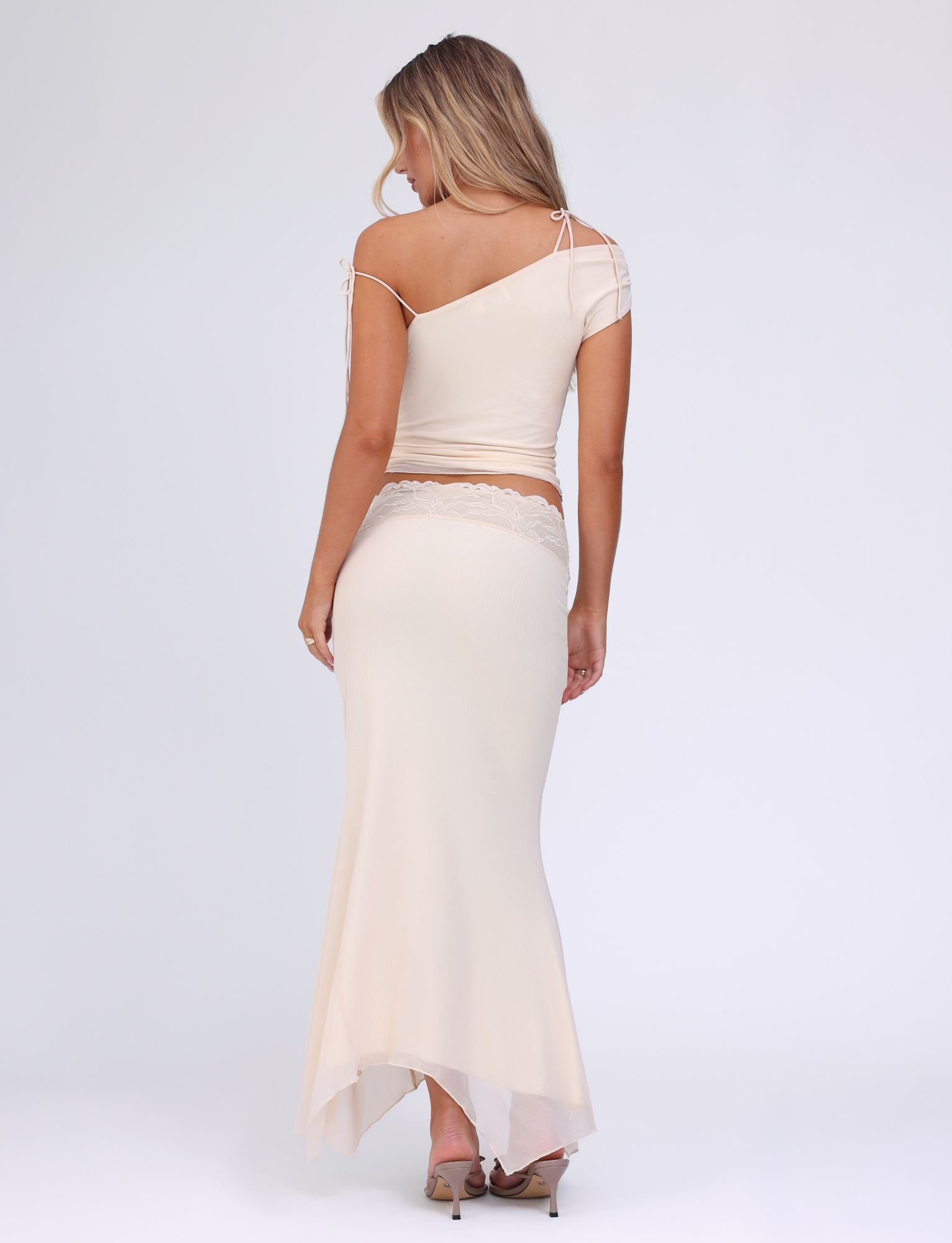 A woman with long blonde hair models the TIGERMIST LILLIAN SKIRT in white, cream, and floral print, featuring an off-the-shoulder design with an asymmetrical hem and mesh fabric. She stands in heels against a plain white background, seen from the back.