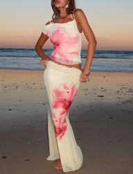 A woman stands barefoot on the beach at sunset, wearing the CL - LILLIAN SKIRT by Tiger Mist USA—a cream outfit with large pink flower prints, sleeveless top, and long skirt. The ocean waves and sunset sky create a picturesque backdrop.