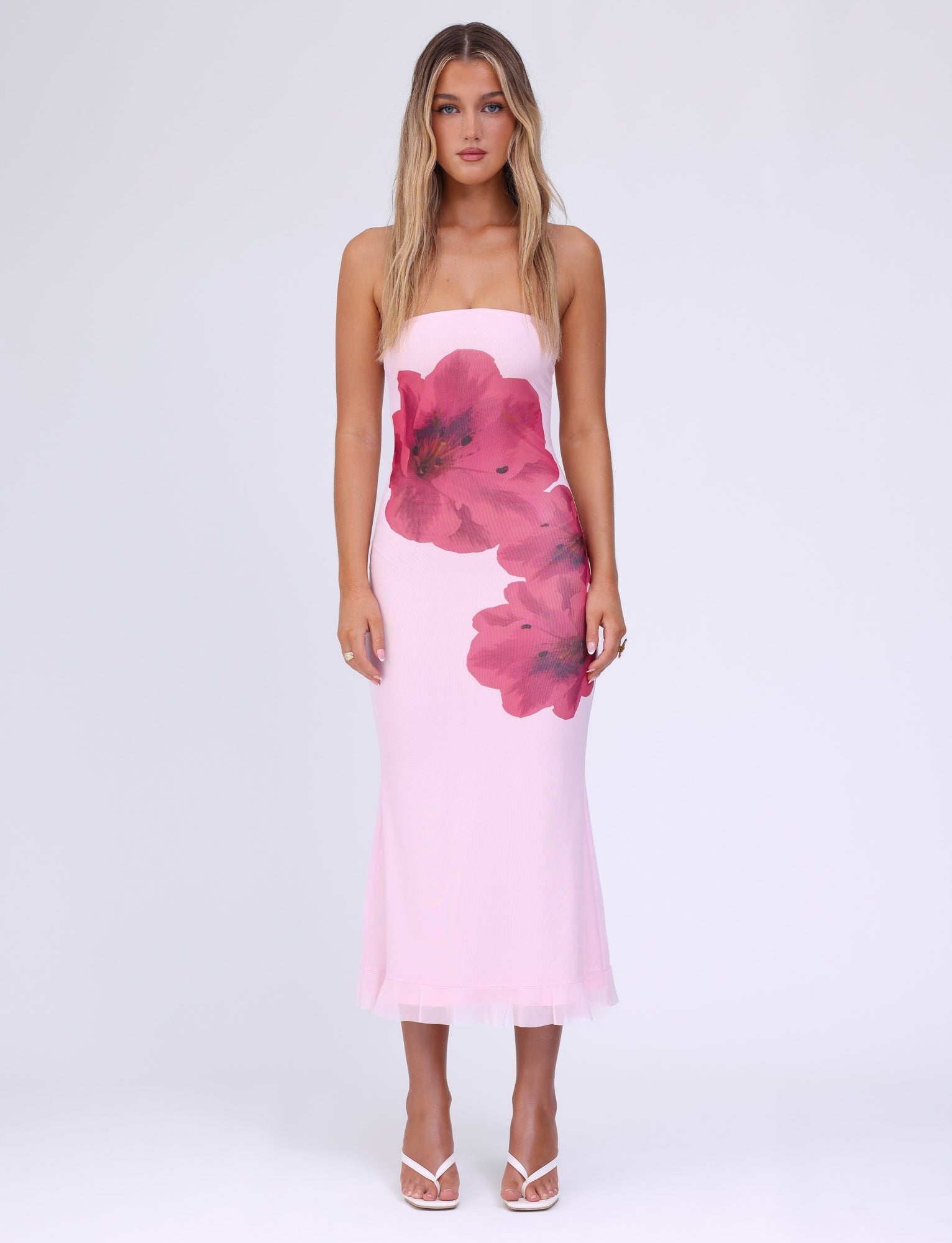 A woman with long, wavy blonde hair is donning the TIGERMIST LILLIAN DRESS in baby pink, a strapless midi-length dress adorned with floral print designs. The dress perfectly complements her white high-heeled sandals as she stands against a plain white background.
