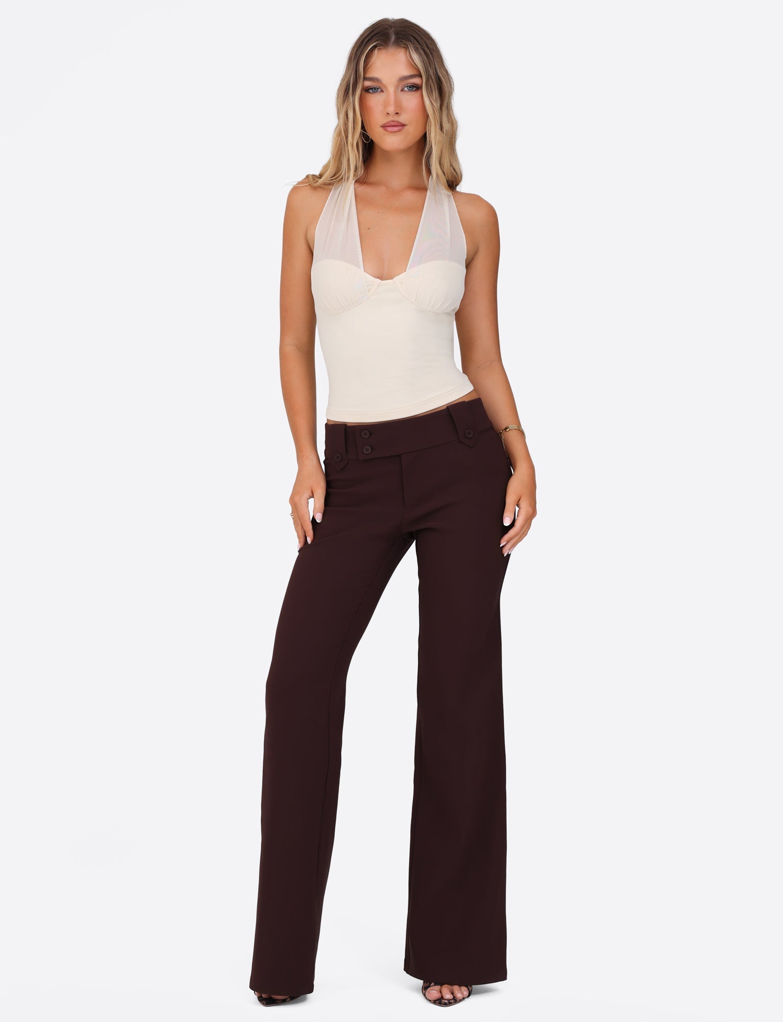 A woman poses against a white background, wearing the TIGERMIST GRANGER TOP in cream, featuring a halter neckline, paired with dark brown wide-leg pants. She stands confidently, with loose hair and hands by her sides.