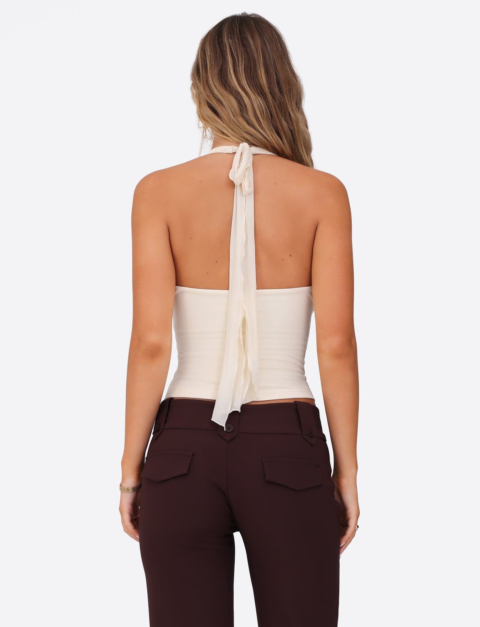 A woman with long hair is seen from behind wearing a white cream GRANGER TOP by TIGERMIST, featuring a halter neckline with a long, flowing tie and dark brown trousers, all against a plain white background.