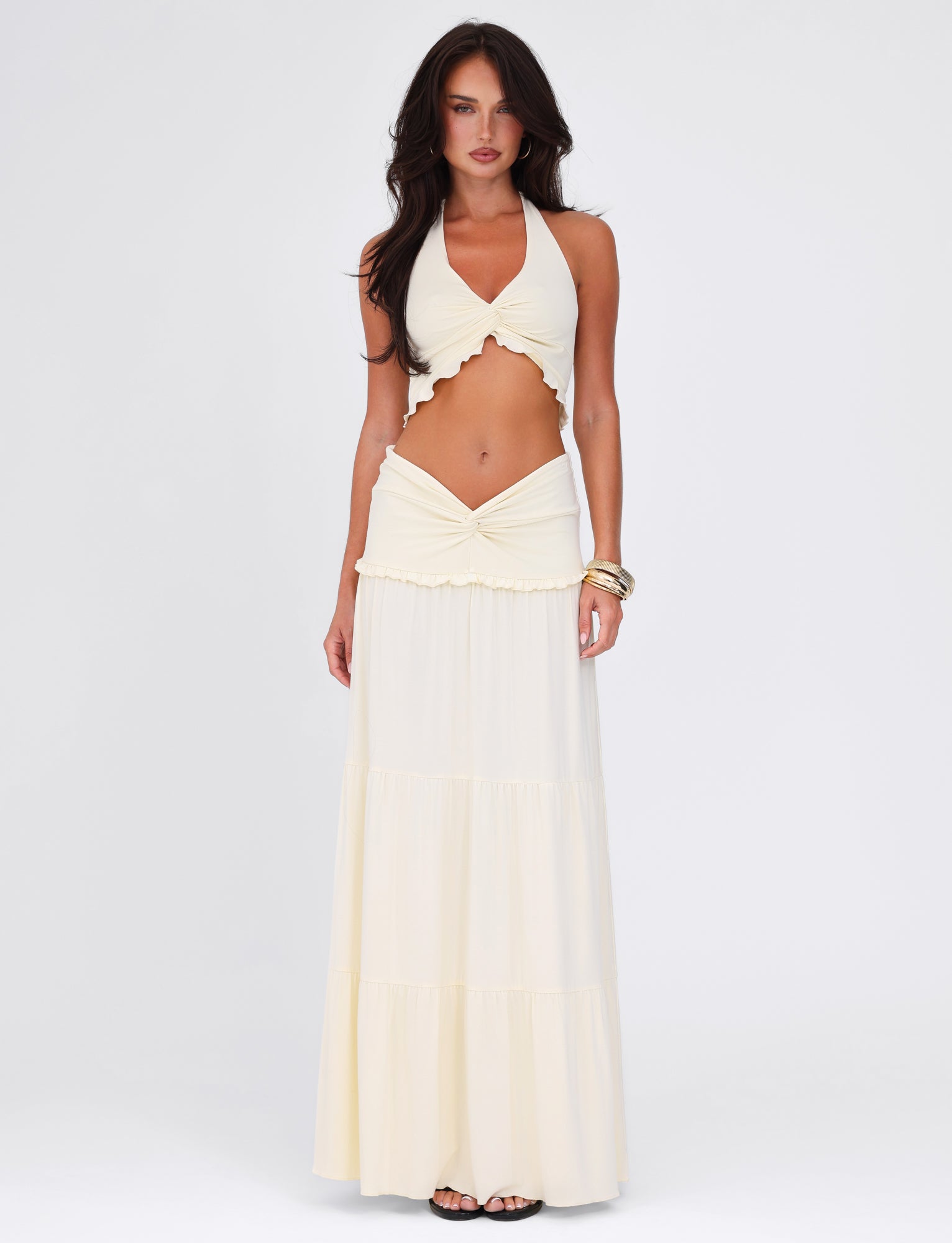 A woman showcases the TIGERMIST KATYA MAXI SKIRT in white and cream. The outfit features a sleeveless high-stretch halter neck top with a twist detail cutout at the midsection and a flowing maxi skirt. She wears bracelets, and her long, dark hair cascades over her shoulders against a white background.