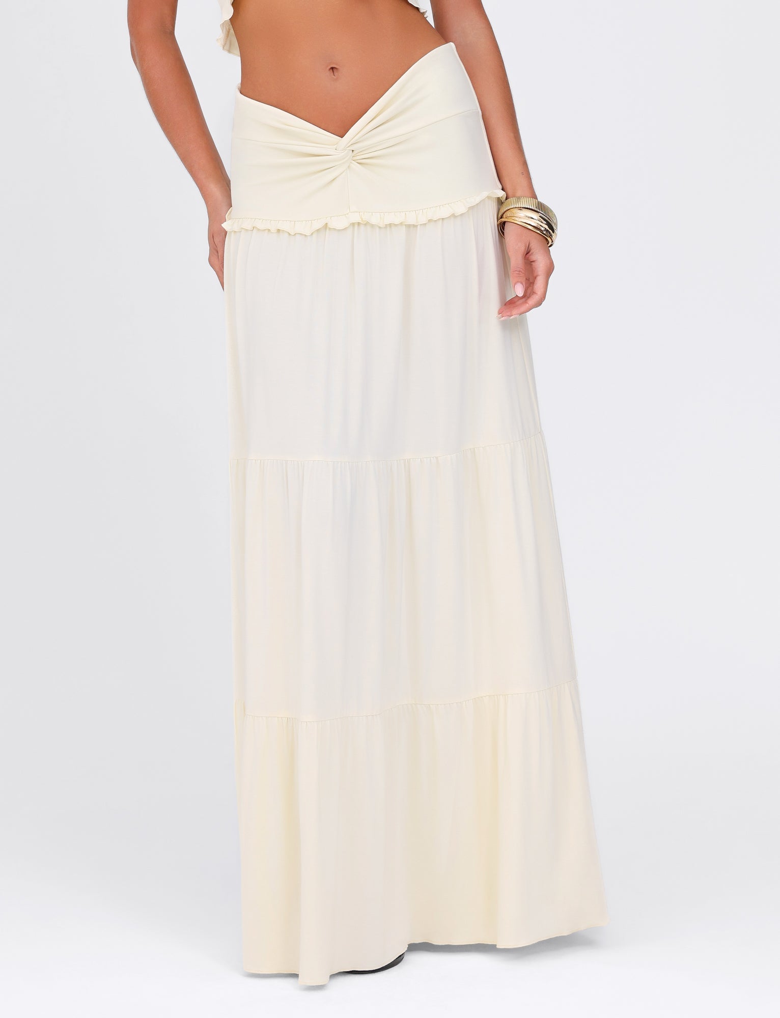 A person wearing the KATYA MAXI SKIRT - WHITE : CREAM by TIGERMIST, featuring a knotted waistband and tiered design for comfort with its high stretch fabric, accessorized with multiple gold bangles against a plain white background.