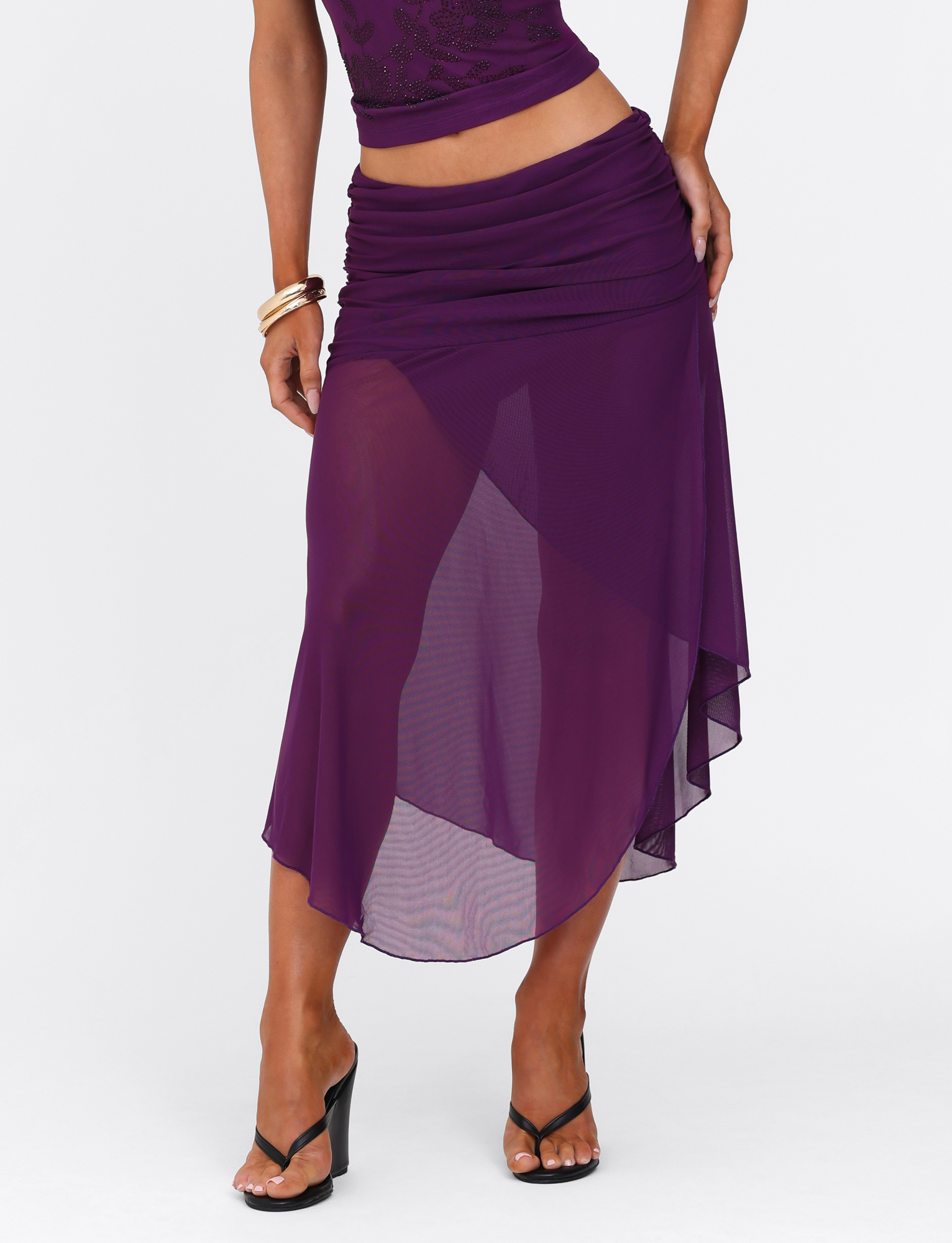 Wearing the TIGERMIST MARIANA SKIRT - PURPLE, a sheer asymmetric wrap skirt with waist ruching, paired with a matching top and black heeled sandals. One hand is on the hip; gold bangles accent the other arm.