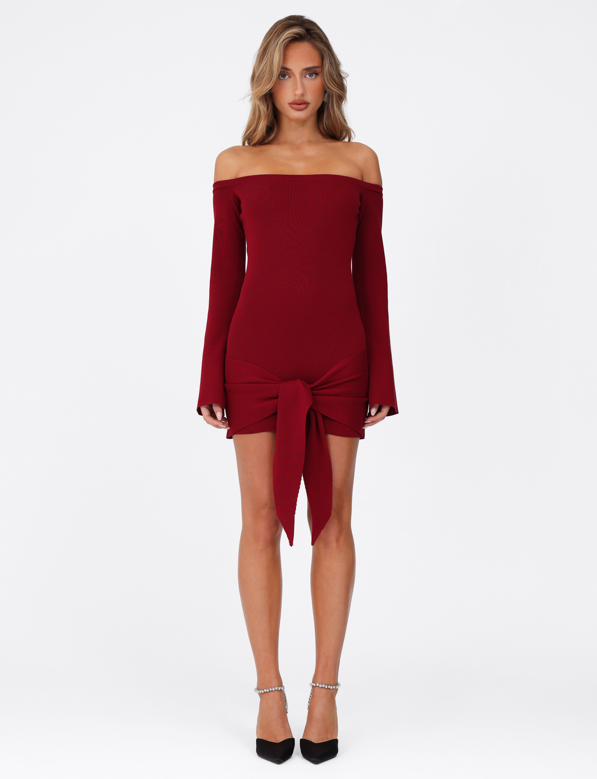 A woman stands against a white background wearing the TIGERMIST CYPRESS MINI DRESS - RED : WINE, a long-sleeve, fitted mini dress with an elastic off-the-shoulder neckline and front tie detail, styled with black pointed-toe ankle strap heels.