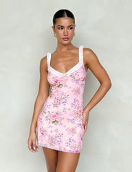 A woman wearing the AMARAN MINI DRESS - PINK : FLORAL : WILD FLOWER by TIGERMIST poses against a plain, light-colored background. Her hair is styled in a sleek bun, and she accessorizes with hoop earrings.