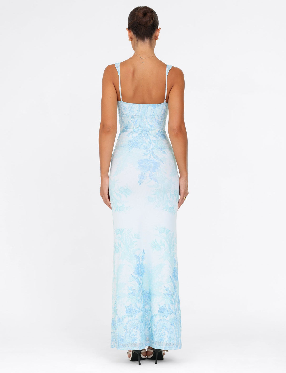 A woman is seen wearing the GUILIANNA FRILL MAXI DRESS from TIGERMIST, featuring a fitted silhouette with a blue floral tapestry pattern and frill shoulder straps. The dress showcases thin straps and a straight neckline. Her hair is styled in an updo, and she pairs the look with black heels against a plain white background.