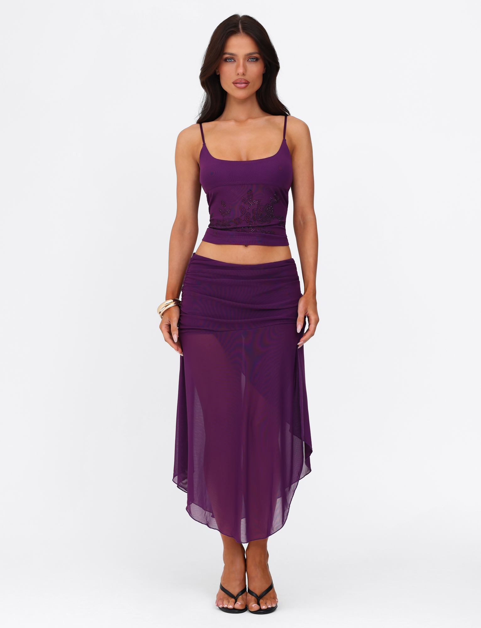 A woman stands against a white background wearing the TIGERMIST OCTAVIA TOP in purple, featuring floral embellishment, paired with black high-heeled sandals and gold bracelets.