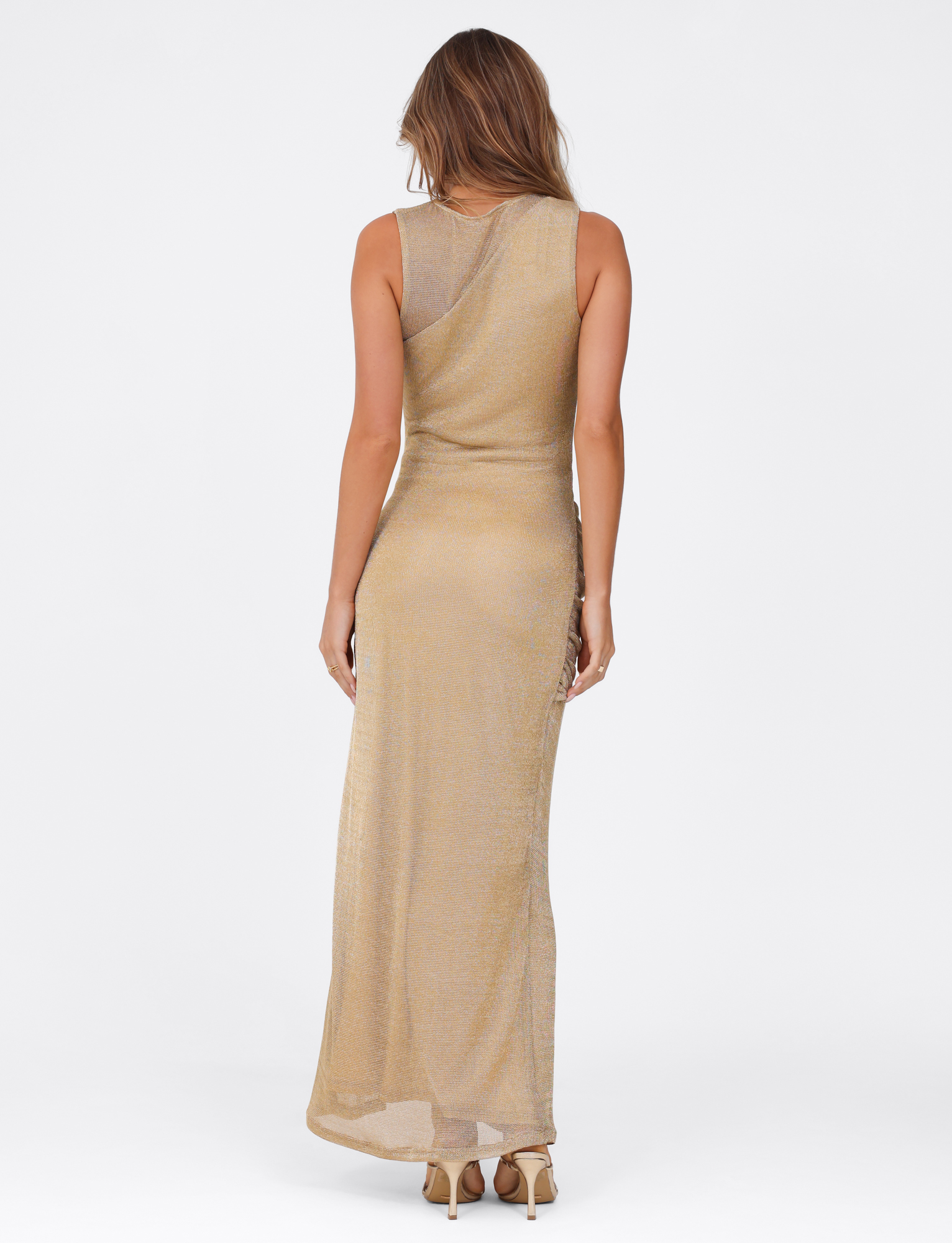 A woman faces away wearing the TIGERMIST AZELMA MAXI DRESS in gold—a sleeveless, floor-length metallic knit dress—paired with beige high-heeled sandals against a plain white background.