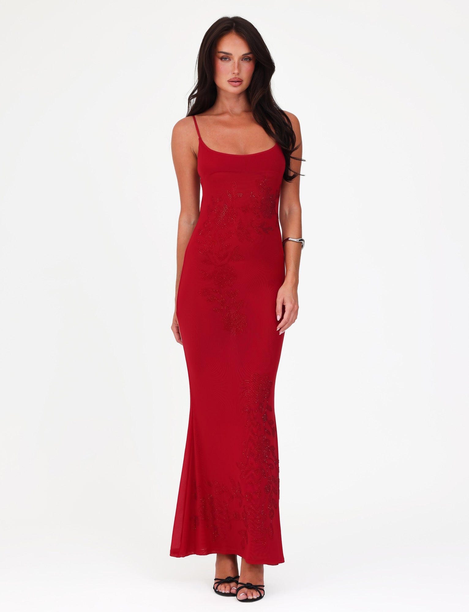 A woman in a sleeveless, cherry red Octavia Maxi Dress by TIGERMIST stands against a white background. She is wearing black high heels and a silver bracelet, with long dark hair cascading over her shoulders. The dress features subtle floral embellishments that add an elegant touch.
