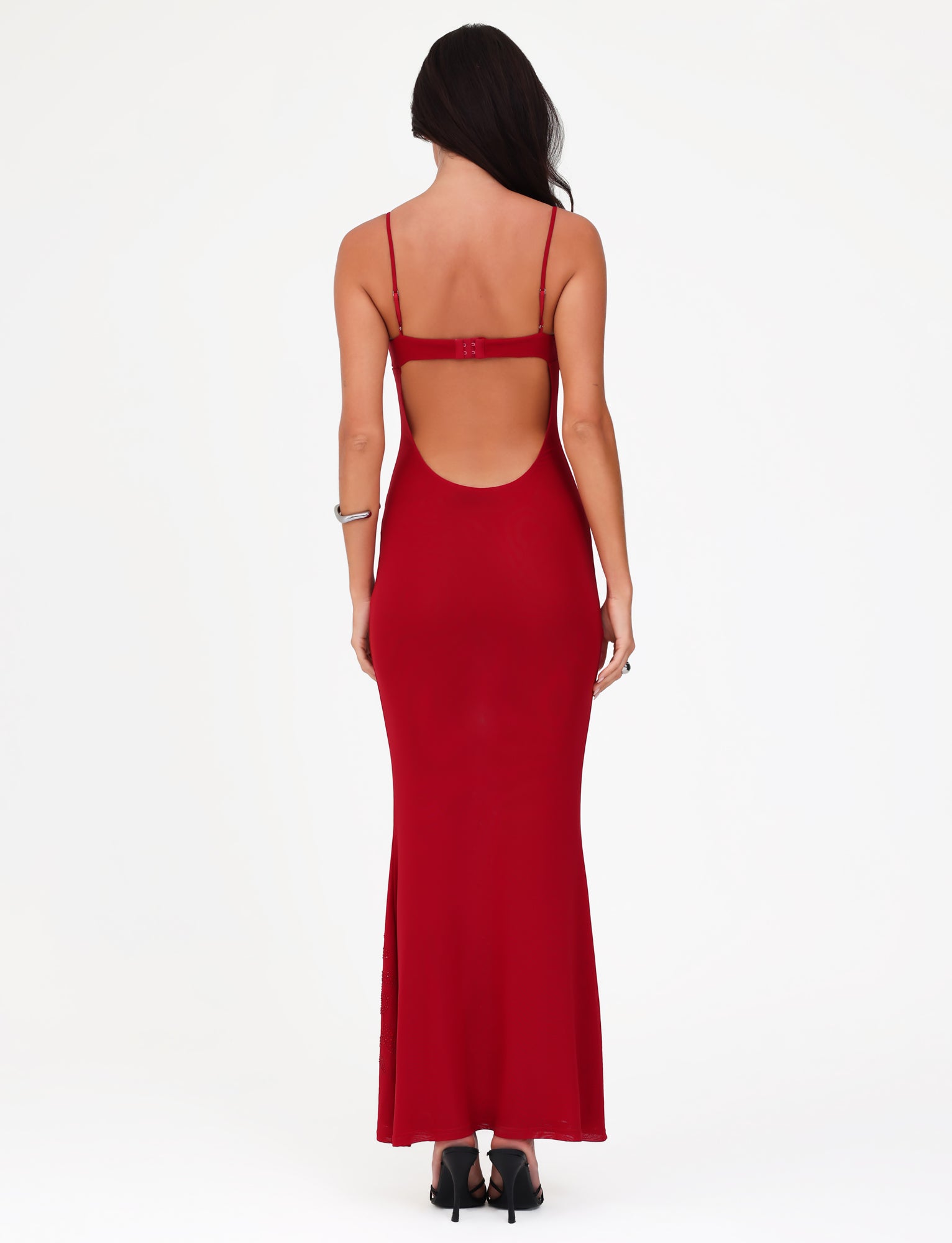 Rear view of a woman in a Tigermist Octavia Maxi Dress in Cherry Red, featuring a backless design with spaghetti straps, standing against a plain white background. The maxi-length dress pairs perfectly with her black heels and the bracelet on her right wrist. Her long hair cascades down her back, accentuating the elegant look.