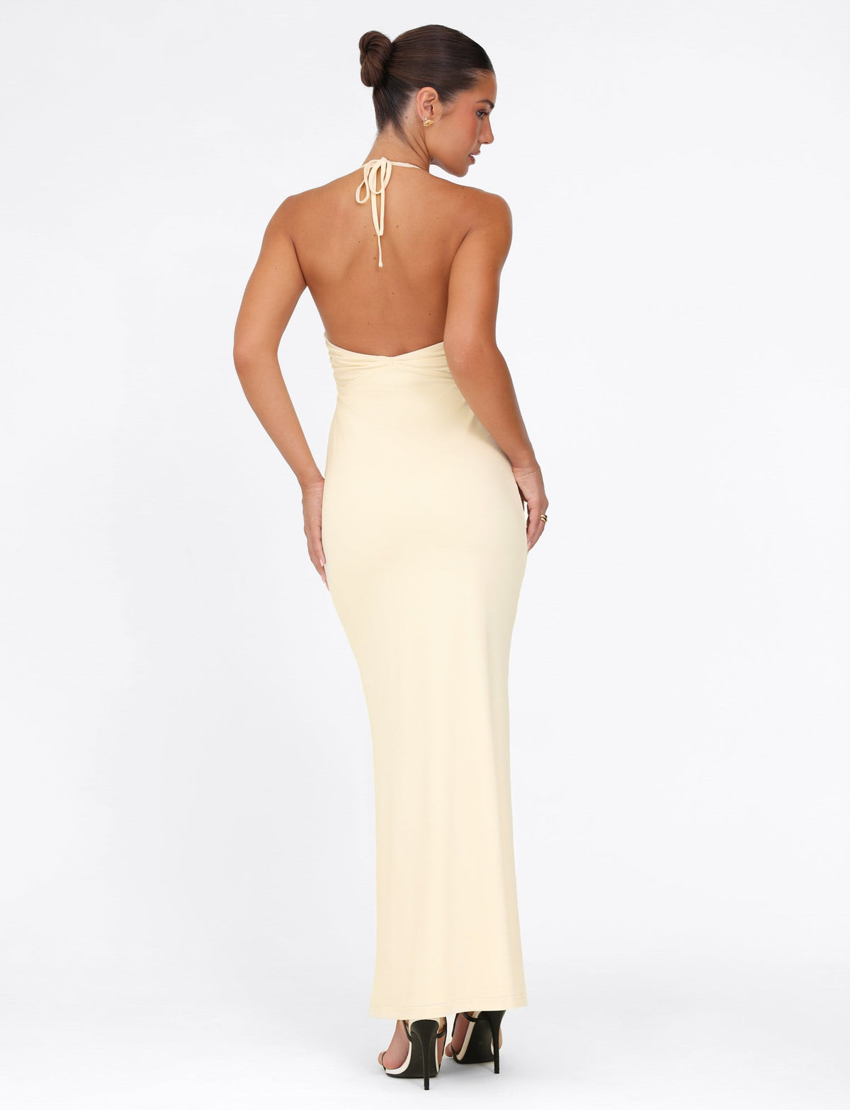 A woman in an elegant lemon Deena Maxi Dress with lace bust cups, designed by TIGERMIST, stands with her back to the camera. Her hair is styled in a neat bun, and she wears black high heels. The dress's maxi length flows gracefully against the plain white background.