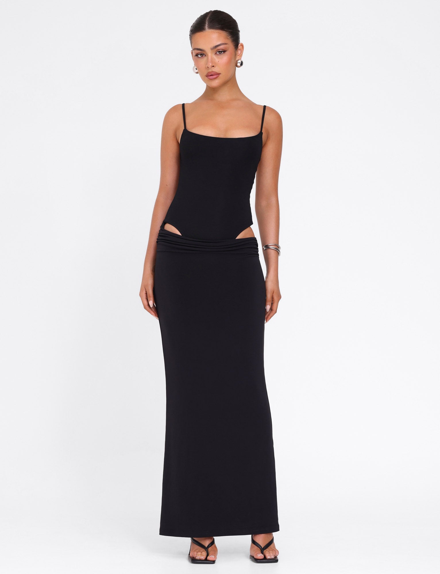 A woman poses against a white background in the TIGERMIST UMA DRESS - BLACK, a bodycon maxi dress featuring adjustable shoulder straps and hip cutouts. She pairs it with black heeled sandals, hoop earrings, and a sleek updo.