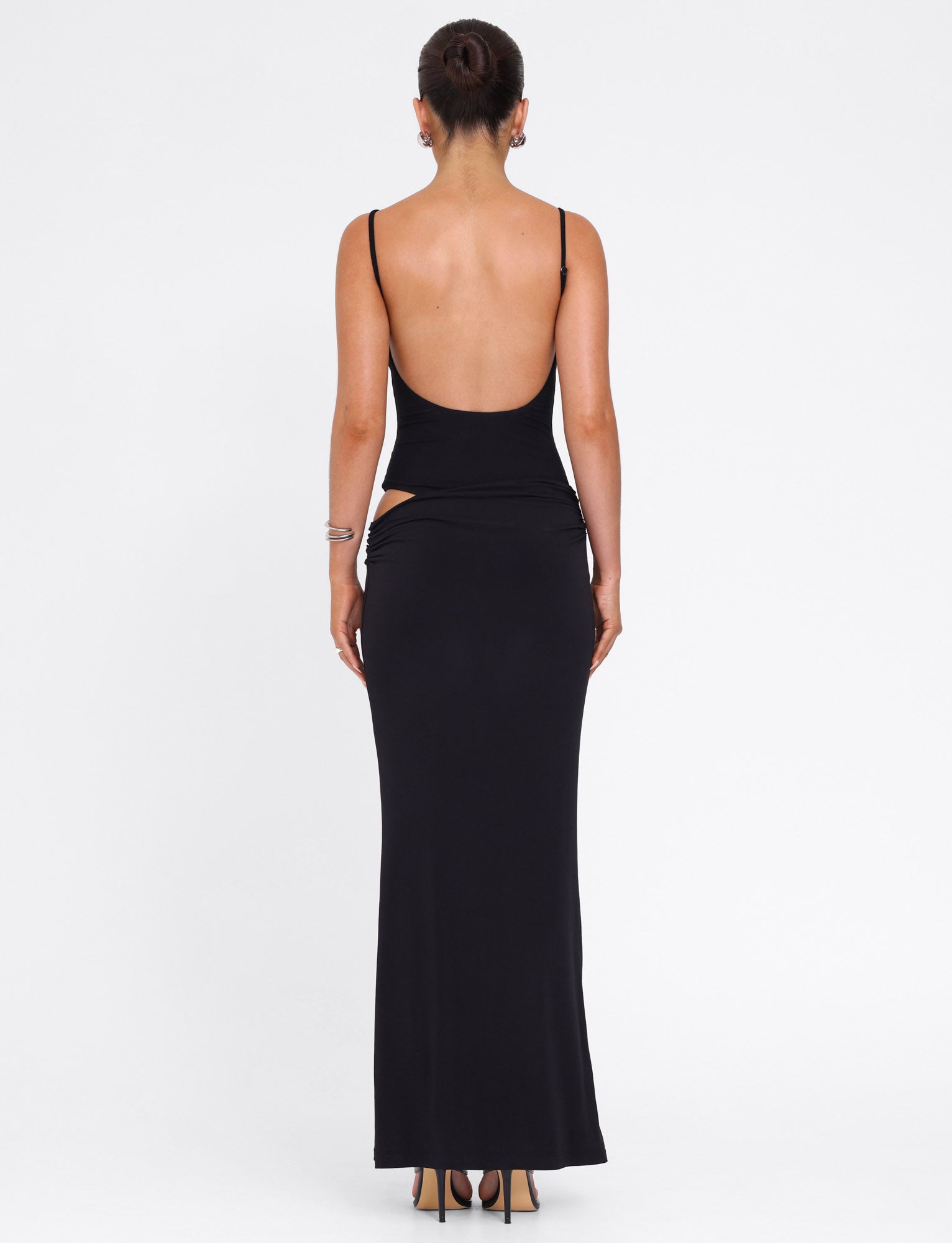 A woman stands facing away in the UMA DRESS - BLACK by TIGERMIST, a black bodycon maxi with adjustable straps and a low back. Her hair is in a bun, paired with heels and minimal jewelry, set against a plain white background.