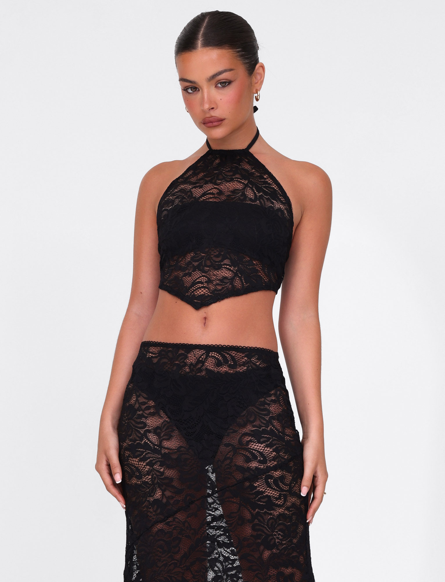 A woman with slicked-back hair and hoop earrings poses against a white background, wearing the TIGERMIST SIYA LACE TOP - BLACK: a sheer black lace high-neck top, paired with a matching high-waisted lace skirt. She has a neutral expression.