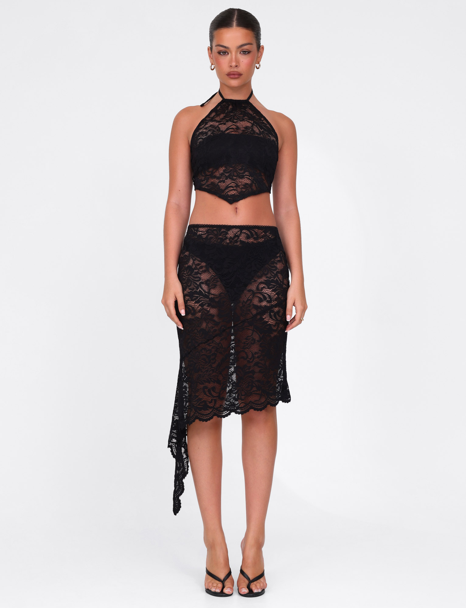 A woman faces forward against a white background, wearing the TIGERMIST SIYA MIDI SKIRT in black lace with an asymmetrical hemline and front slit, styled with a matching black lace halter crop top and black heeled sandals.