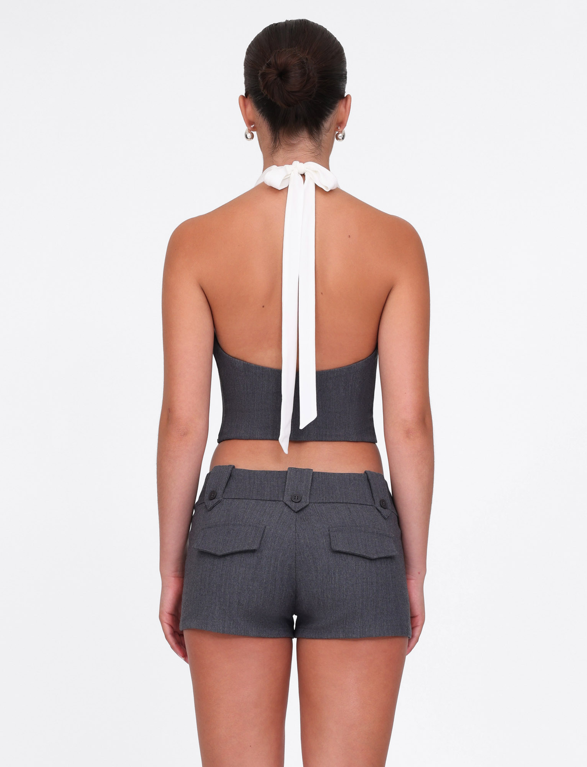 A woman with her hair in a neat bun faces away, wearing the TIGERMIST LOTUS TOP in cream-grey textured jersey and matching high-waisted shorts with a stretch suiting waist and buttoned back pockets, set against a plain white background.