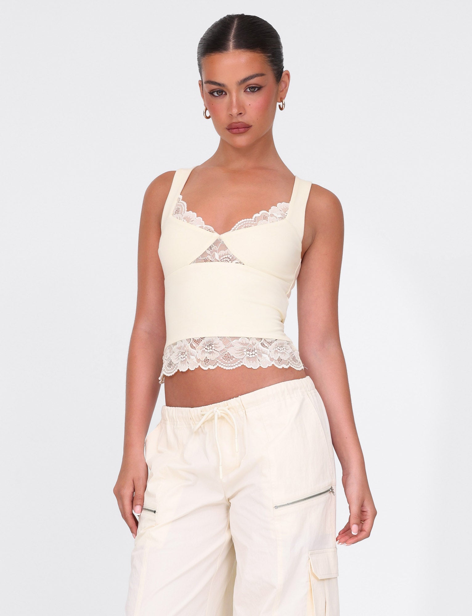 A woman with dark hair pulled back wears the Dione Lace Top in soft butter by Tigermist and off-white cargo pants, standing against a plain white background.