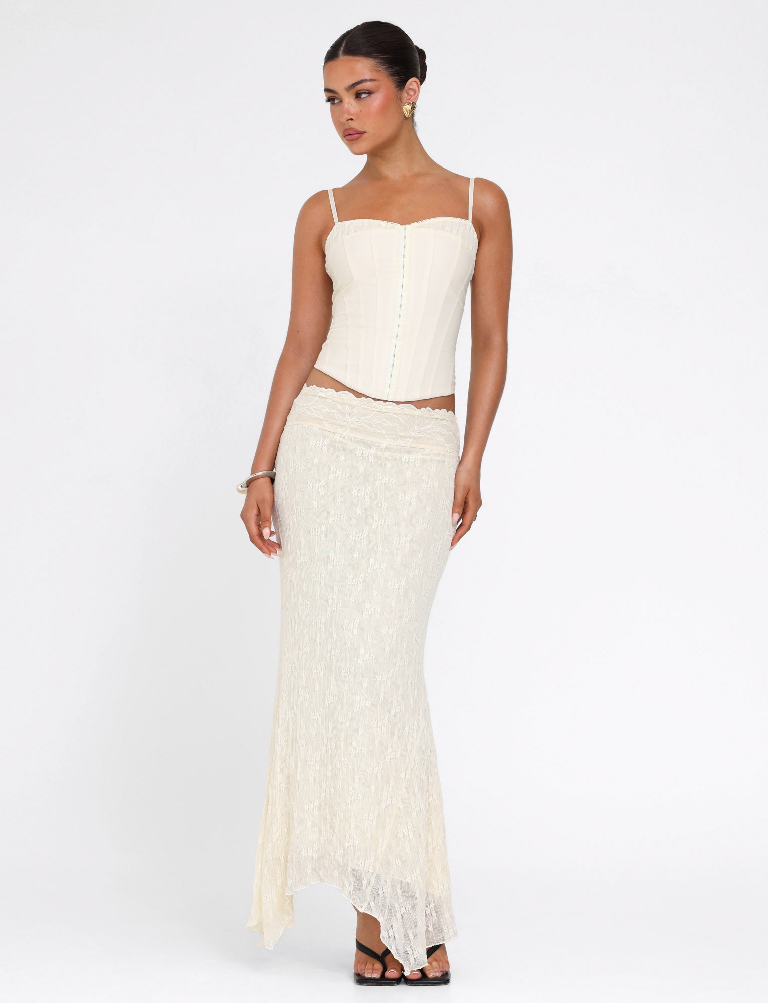 A woman stands against a white background wearing the TIGERMIST ANTONELLA TOP in cream with thin straps, paired with a matching long suiting skirt. Her hair is styled back and she completes the look with black heeled sandals.