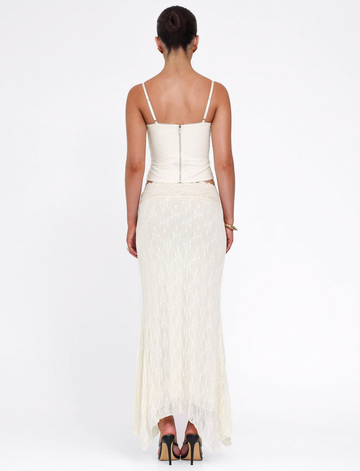 A woman with her hair in a bun is seen from behind, wearing a sleeveless white top and the TIGERMIST ANTONELLA MIDI SKIRT in cream lace, paired with high heels, standing against a plain white background.