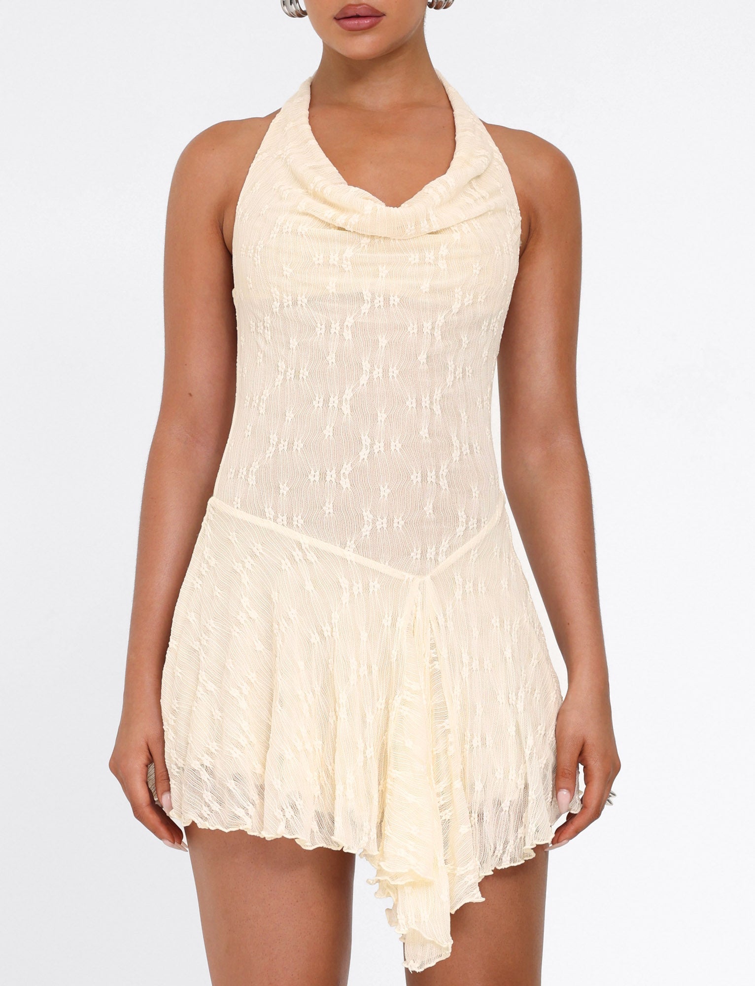 A woman stands against a plain white background wearing the TIGERMIST FIORELLA MINI DRESS in cream lace, featuring a sleeveless halter neck, draped neckline, and asymmetrical hem.