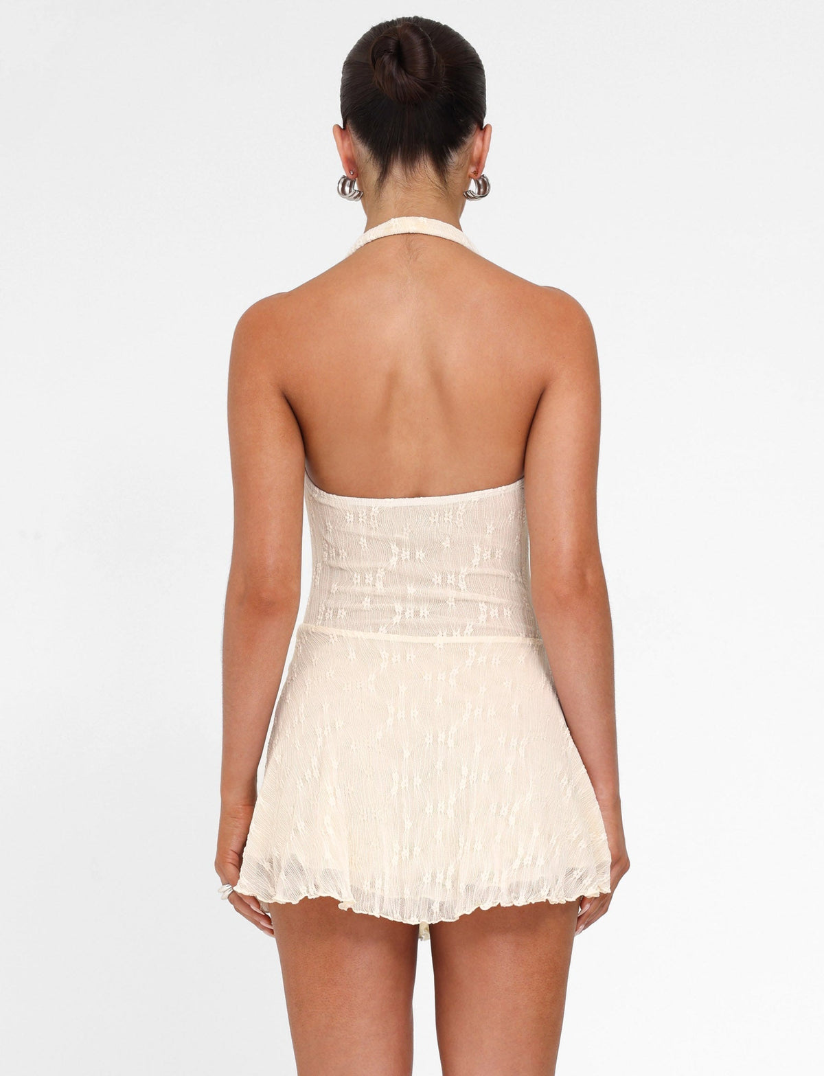 A woman with her hair in a bun and hoop earrings models the TIGERMIST FIORELLA MINI DRESS in cream lace, featuring a halter neck, fitted bodice, and flared short skirt, against a plain white background.