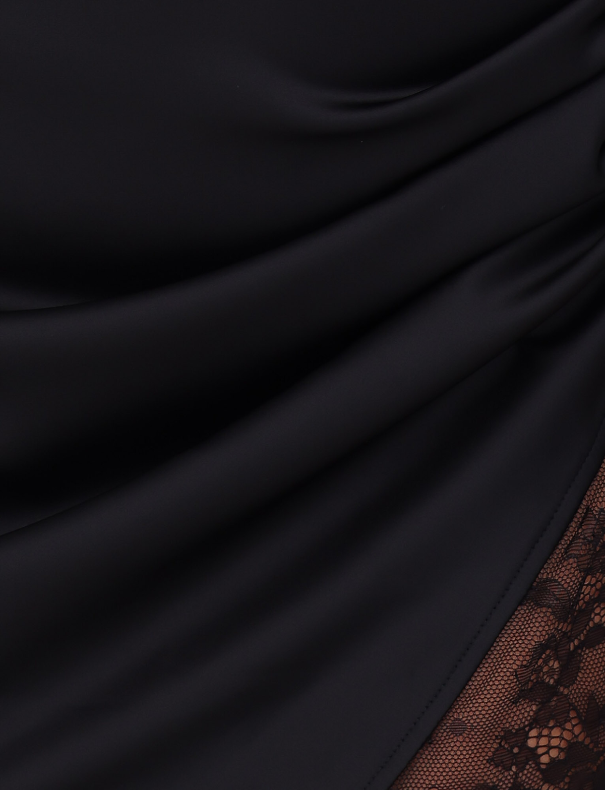 Close-up of the TIGERMIST BEXLEY MINI DRESS in black, showing smooth satin fabric with soft folds and a lace panel overlay on a sheer brown background in the lower right corner.