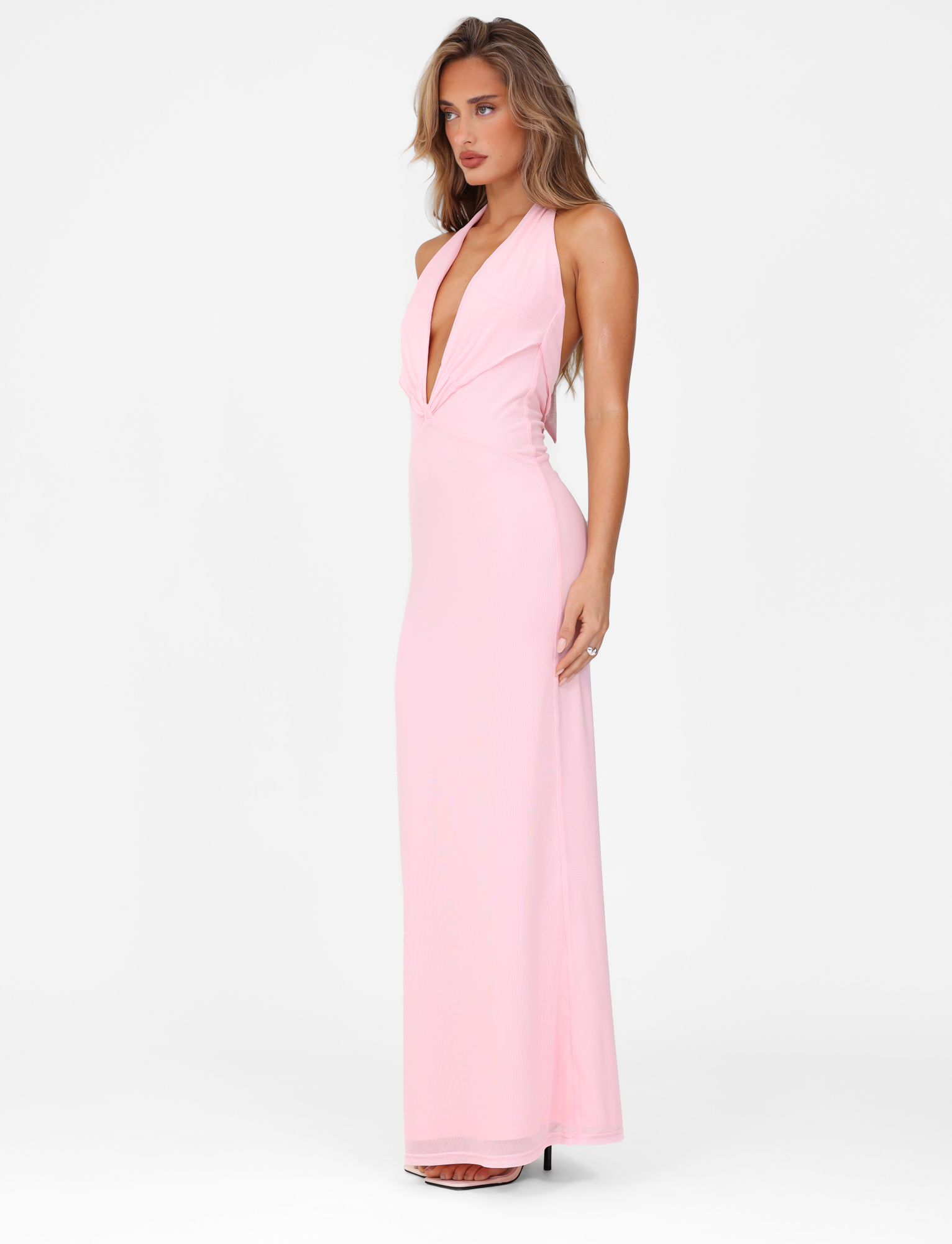 A woman poses against a white background in the TIGERMIST CRESSIDA MAXI DRESS - PINK: BABY PINK, a sleeveless, maxi-length dress with a plunging neckline and high heels. She stands slightly turned with a relaxed expression.