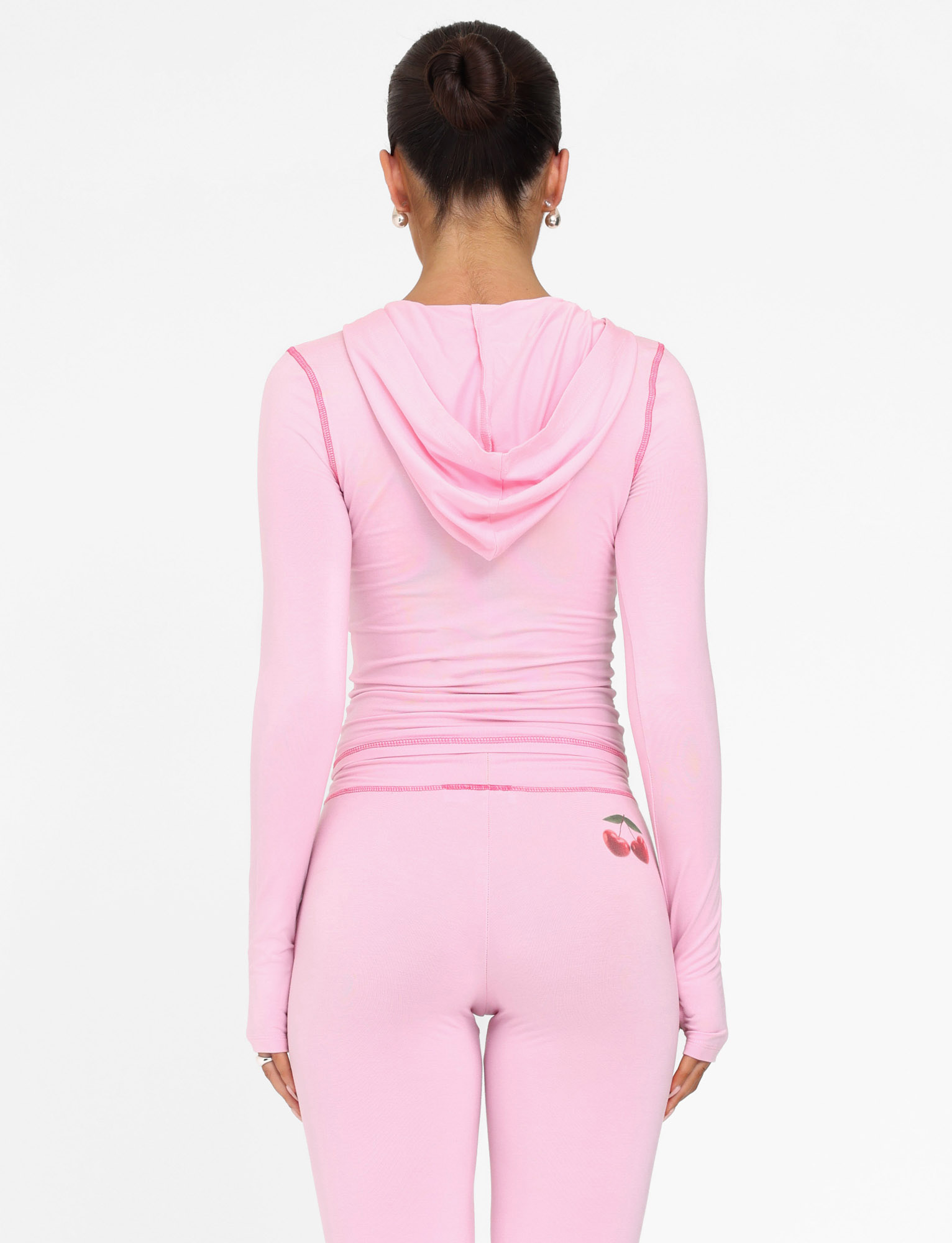 A dark-haired woman in a bun wears the TIGERMIST BLAYZE HOODIE - PINK : SOFT PINK and matching leggings with a cherry graphic, facing away against a plain white background.
