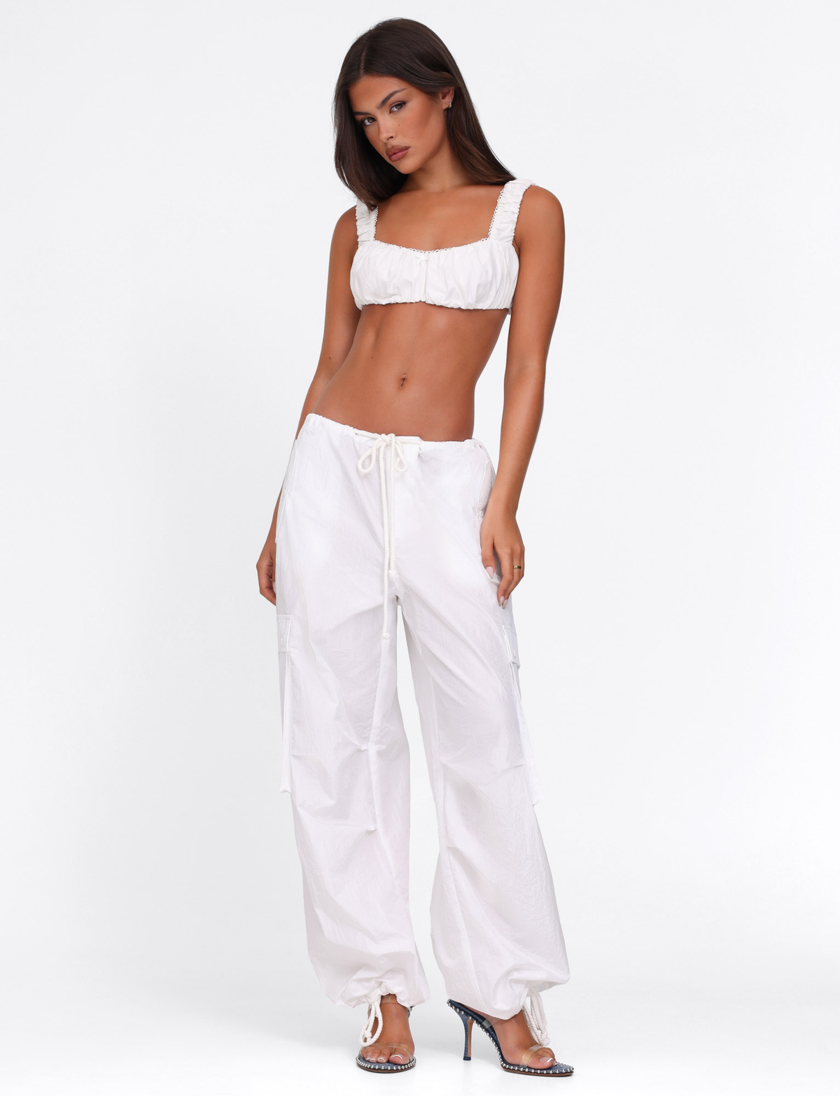 A woman poses against a white background wearing the TIGERMIST SABRA TOP in white, paired with loose drawstring pants and silver high-heeled shoes.