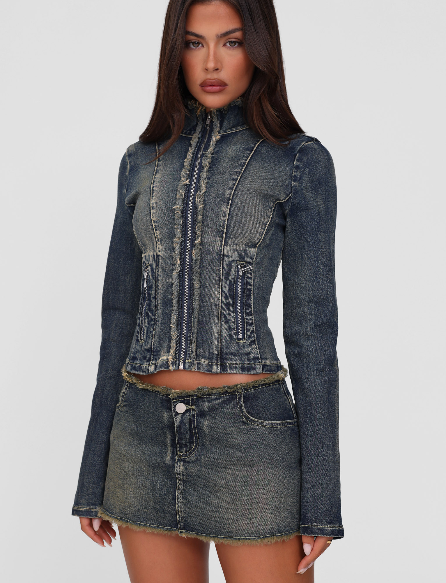 A woman wears the TIGERMIST OPHELIE JACKET in denim blue, featuring a vintage wash, frayed edges, long sleeves, and vertical seams for a distressed look. She pairs it with a matching mini skirt against a plain light background.