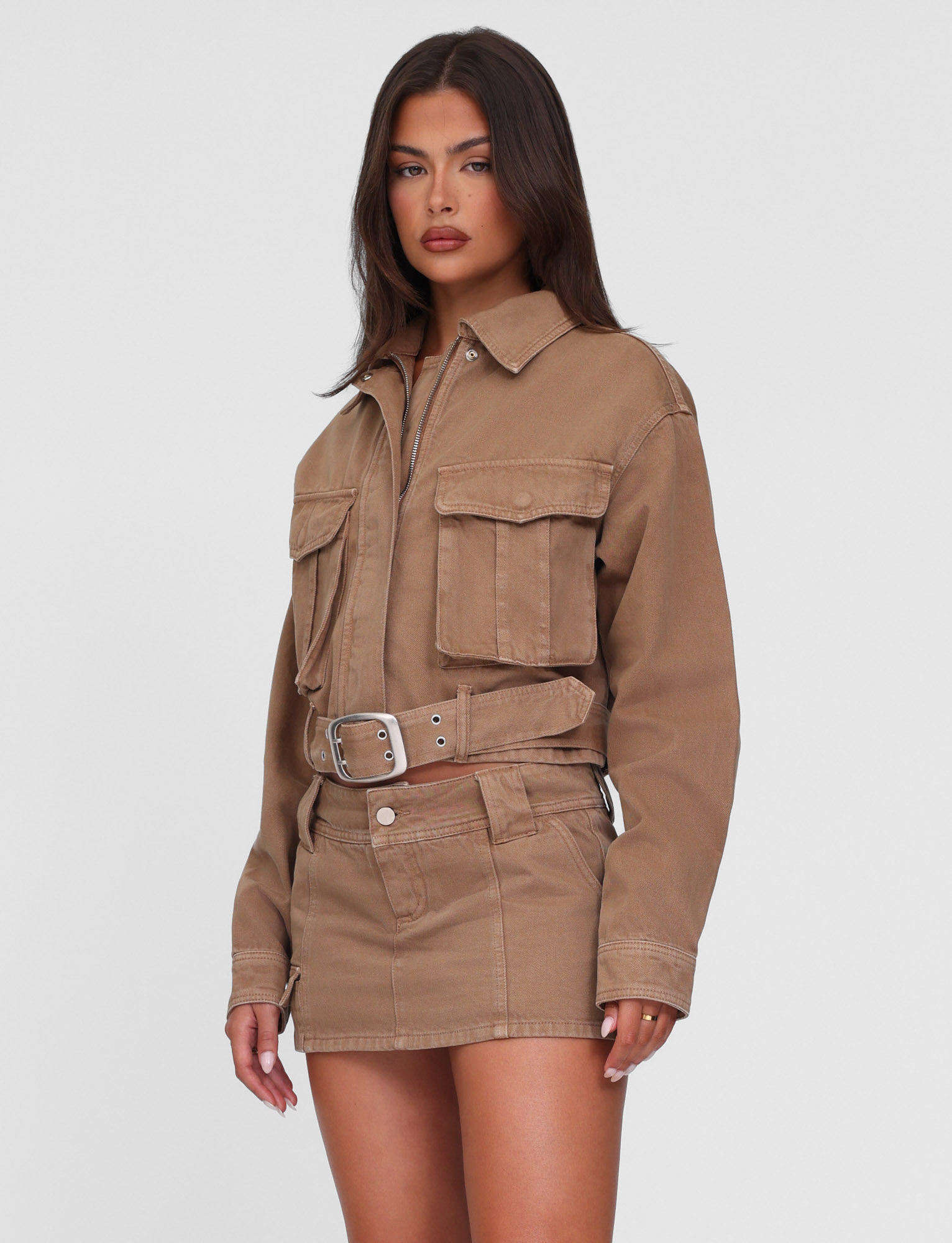 A woman with long dark hair and a neutral expression poses against a white background, wearing the TIGERMIST ZELIE MINI SKORT in washed tan and a matching tan denim jacket with functional pockets.