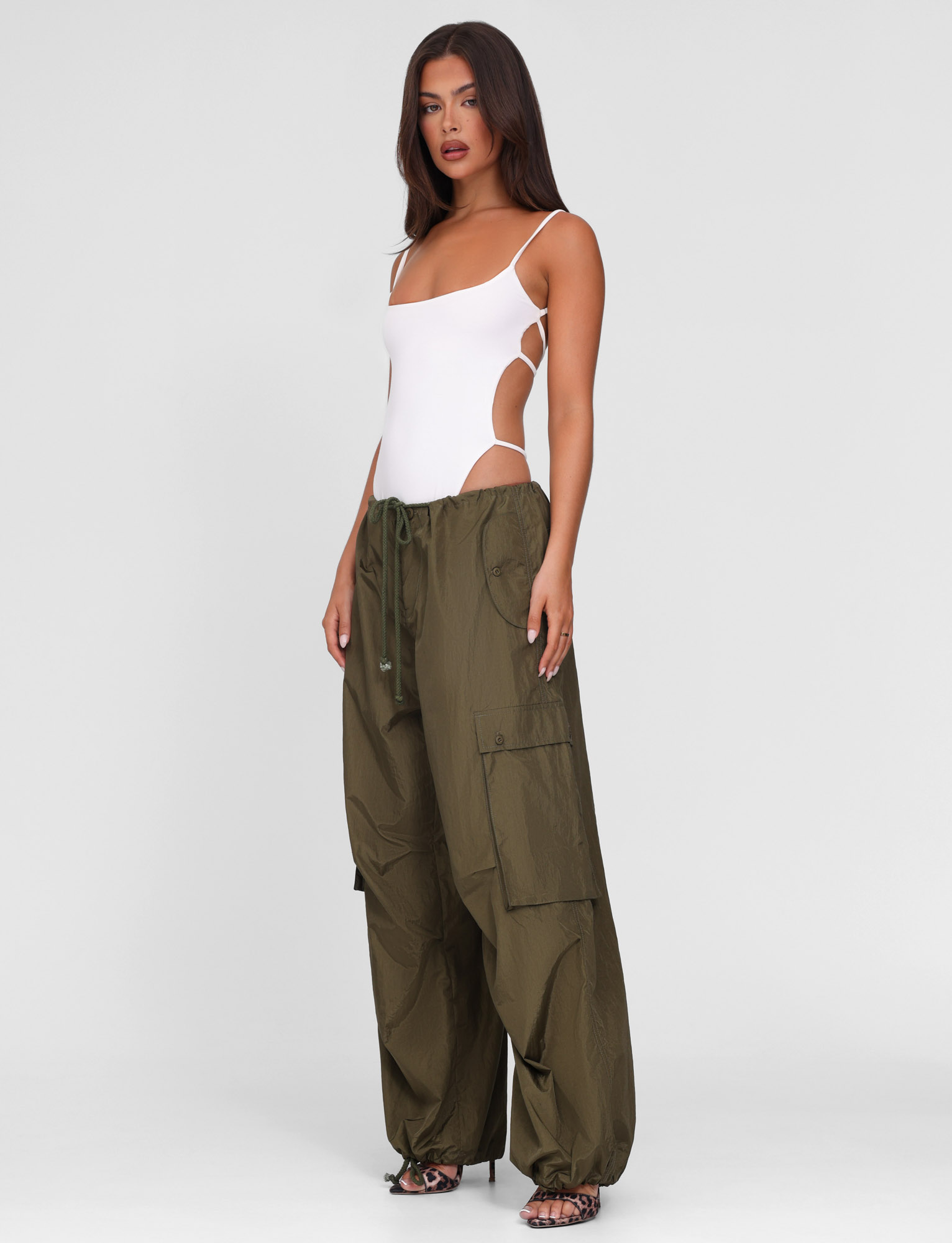 A woman poses against a plain background wearing TIGERMIST's VIOLETTE PANT in khaki green—loose-fitting cargo pants with an adjustable drawcord waist—styled with a white strappy bodysuit and heeled sandals.