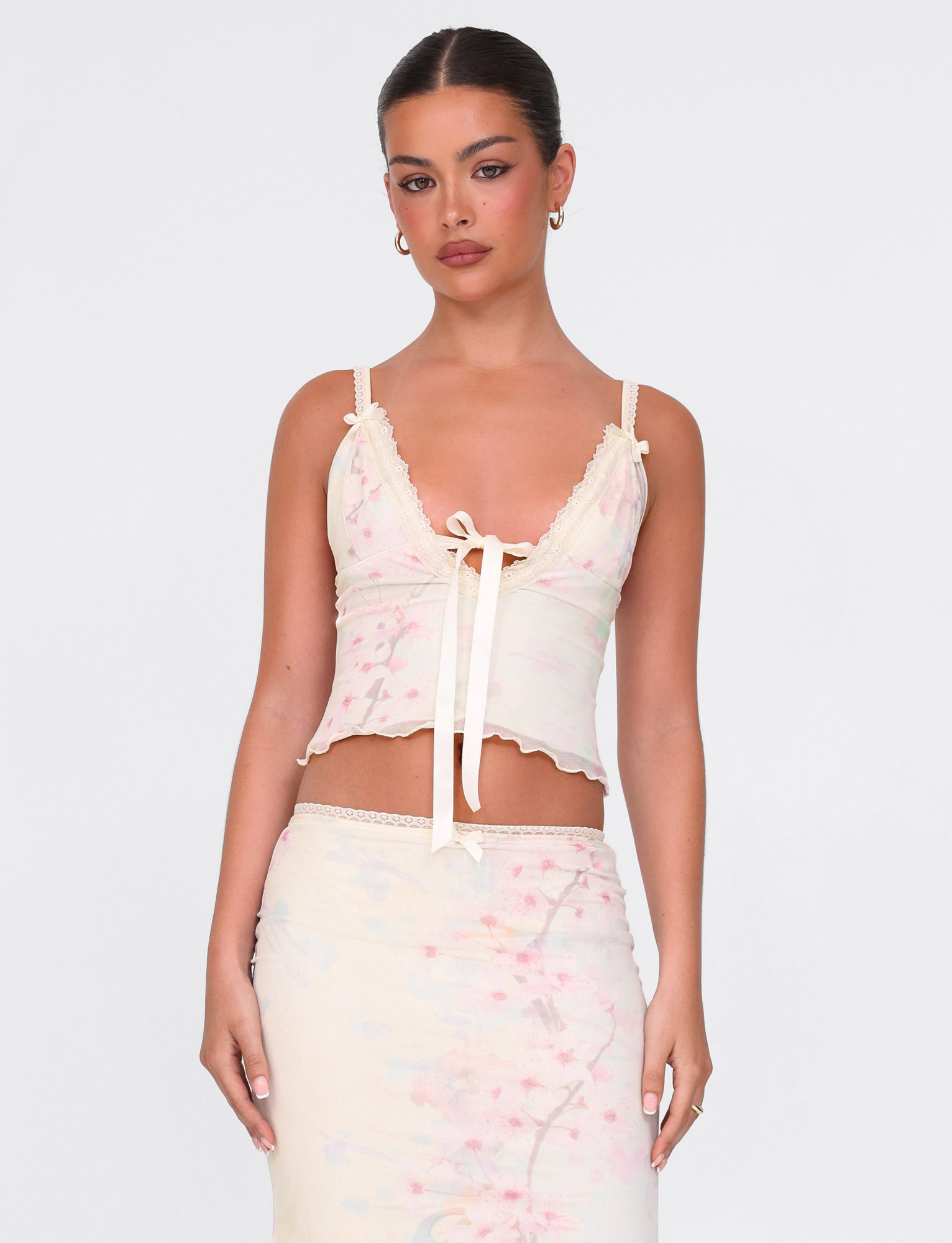 A woman wears the TIGERMIST LONIE TOP in washed cherry blossom floral print, featuring lace trim and a bow, paired with a matching skirt, standing against a plain white background.