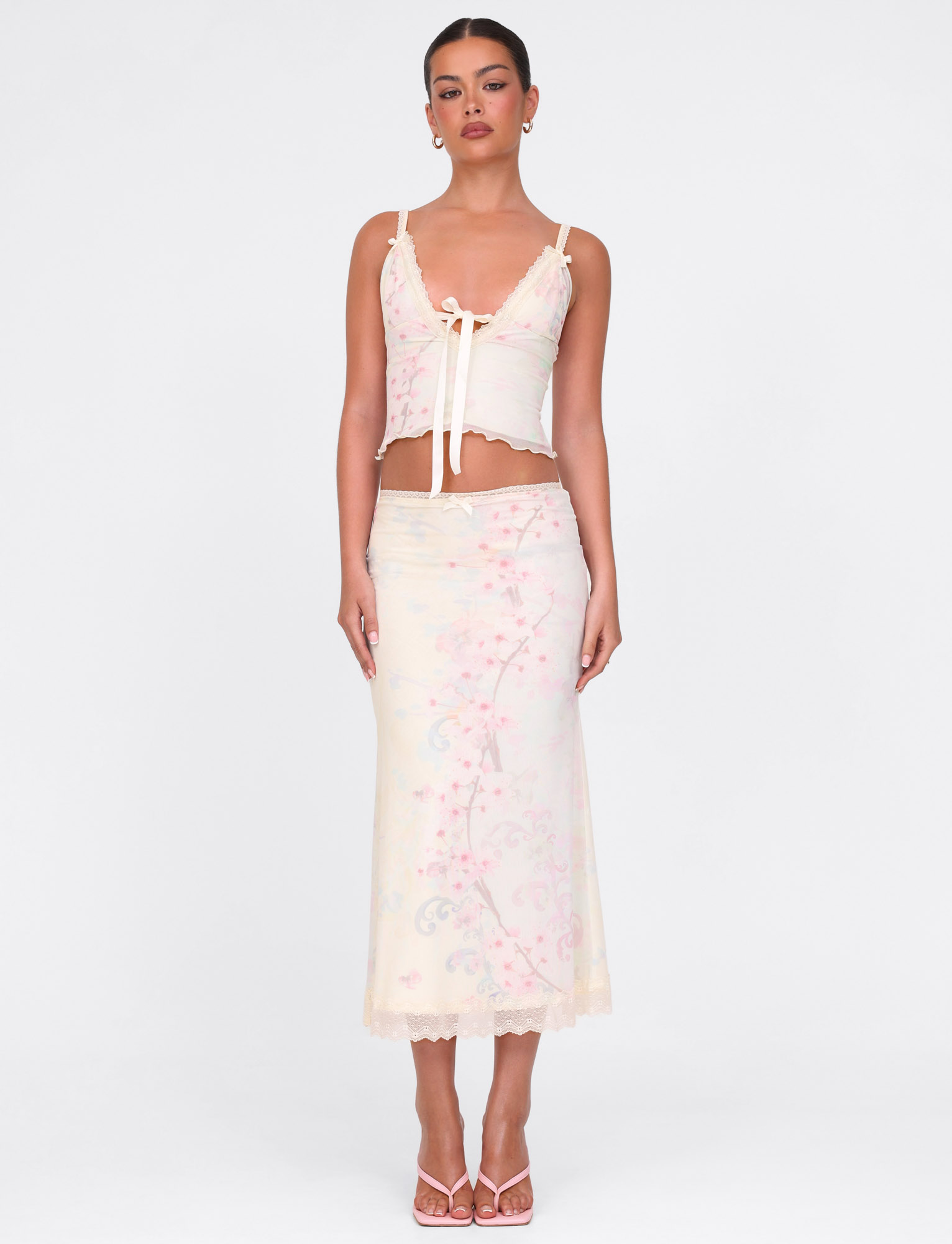 A woman with her hair tied back poses against a white background wearing the TIGERMIST LONIE MIDI SKIRT in Neutral Floral Washed Cherry Blossom, paired with a fitted lace-trim top and strappy heels.