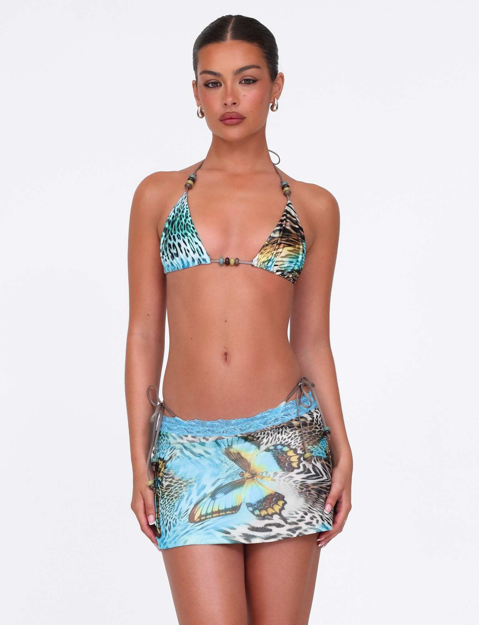 A woman wears the TIGERMIST FINLEA MINI SKIRT - MULTI: ANIMAL: BUTTERFLY ANIMAL and a blue animal print bikini top, standing against a white background and looking at the camera with a neutral expression.