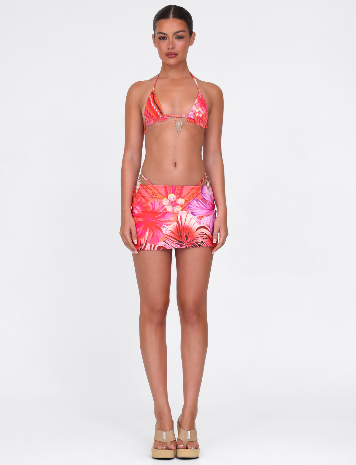 A woman faces forward in the TIGERMIST CIANA BIKINI 2PC BOTTOM - PINK : FLORAL : PINK TROPICAL, featuring a matching mini swim skirt and beige platform sandals, set against a plain white background.
