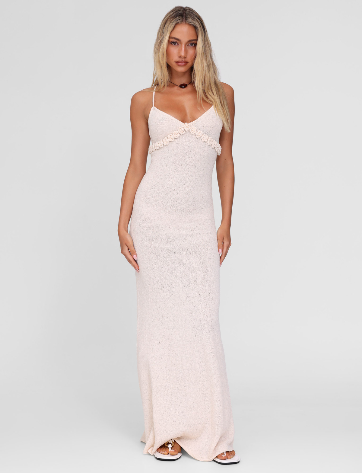 A woman with long blonde hair models the TIGERMIST LAVELLE MAXI DRESS in cream, a light pink, sleeveless, floor-length gown with floral detail and adjustable crisscross straps, standing against a plain white background.