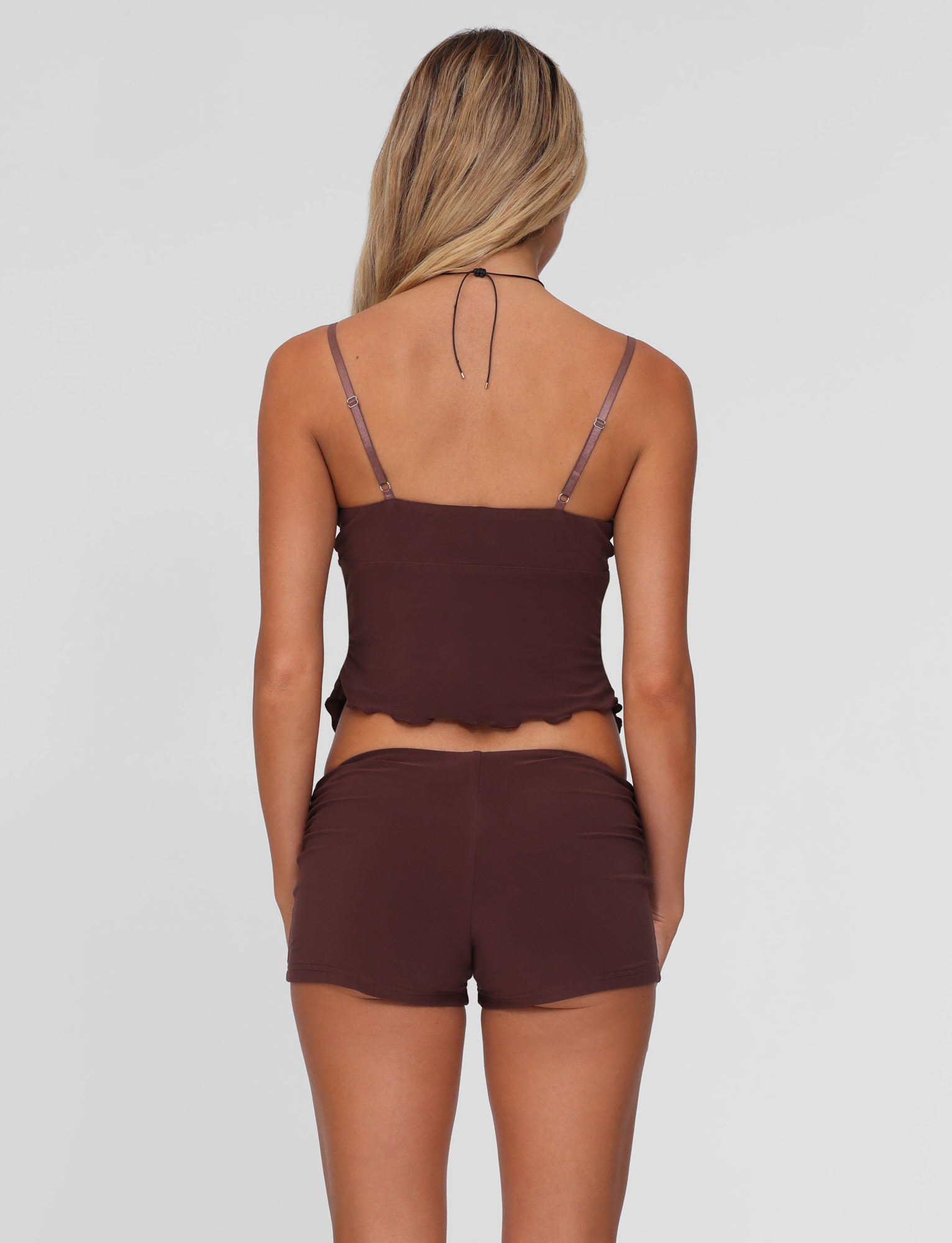 A woman with long blonde hair, facing away, models the TIGERMIST JOVIENNE SHORT - BROWN : CHOCOLATE set—featuring a chocolate brown jersey camisole with spaghetti straps and matching ruched micro mini shorts—against a plain light gray background.
