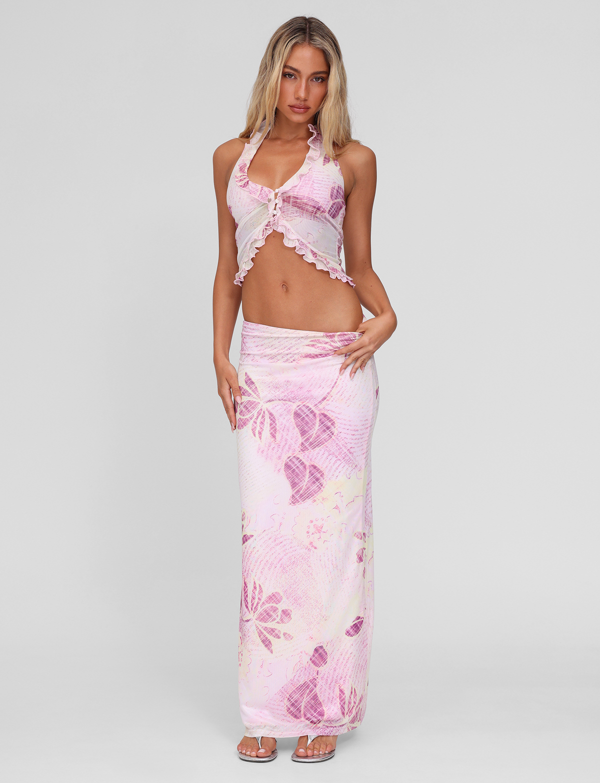 A woman with long blonde hair wears the TIGERMIST DALILA MIDI SKIRT in Grunge Floral Magenta with a matching pink and white floral halter crop top against a plain background, paired with sandals.