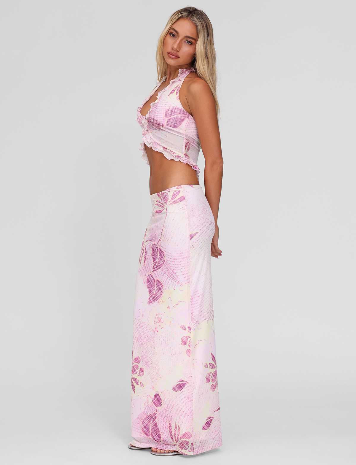 A woman with long blonde hair and neutral makeup poses sideways against a plain light background, wearing TIGERMIST's DALILA MIDI SKIRT in Grunge Floral Magenta with a matching sleeveless pink and white ruffled crop top.