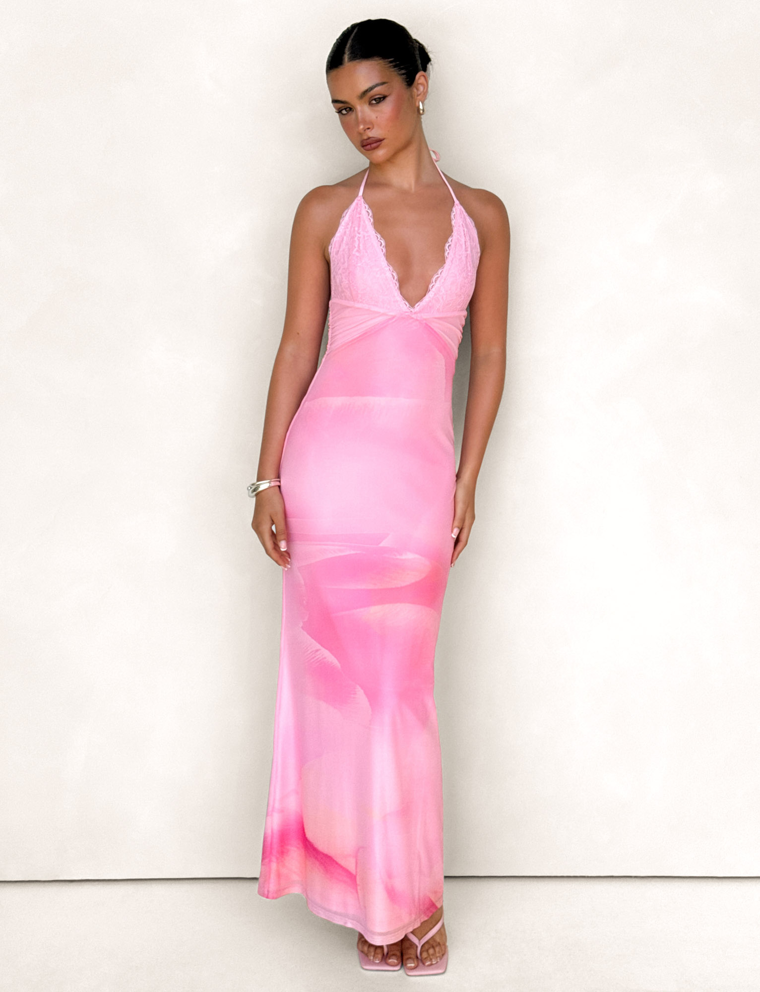 A woman stands against a light background wearing the TIGERMIST DENAE Maxi Dress in Pink Blossom, featuring floral lace and detailed neckline. Her hair is pulled back, and she finishes the look with a bracelet and strappy pink heels.