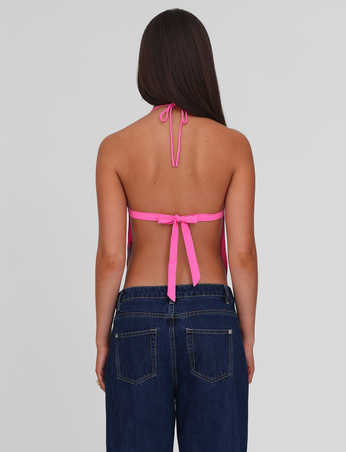 A woman with long brown hair is seen from the back wearing the TIGERMIST KAELAH TOP in hot pink, an open-back halter style tied at the neck and back, paired with dark blue jeans. The background is plain light gray.