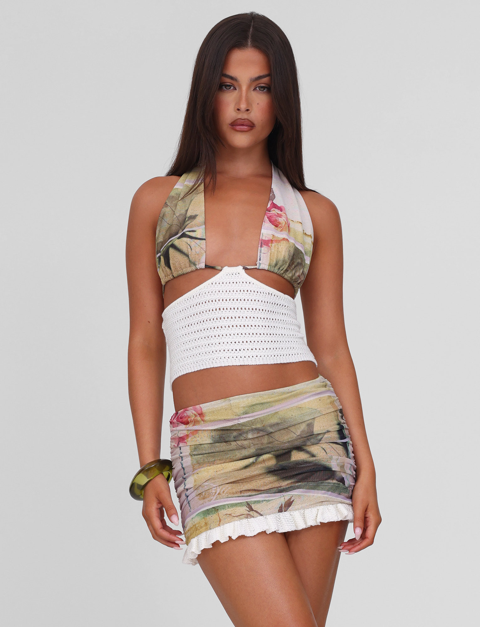 A woman with long brown hair wears the TIGERMIST TILLEY MINI SKIRT - ROSE POSTCARD and a matching floral halter crop top, featuring ruffles and a white crochet band. She stands against a light gray background, looking confidently at the camera.