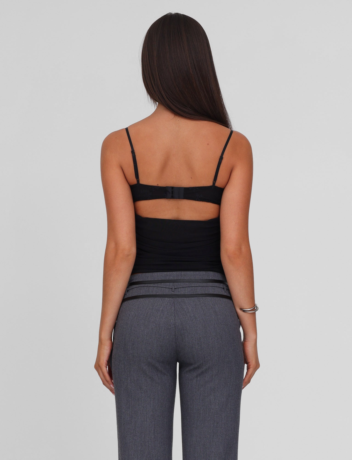 A woman with long straight brown hair, wearing the TIGERMIST KALLIE TOP - BLACK with adjustable straps and gray pants, stands facing away from the camera against a plain light background.