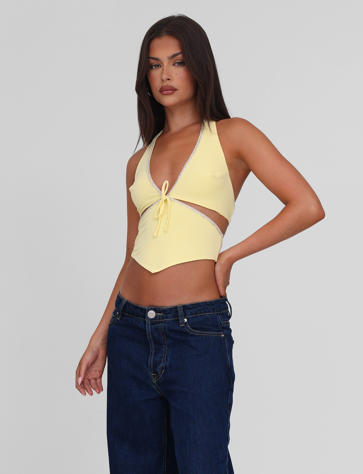 A woman with long brown hair models the TIGERMIST LIZZIE TOP - LEMON, a yellow halter top with cut out details and beaded trim, paired with dark blue jeans against a plain light gray background.