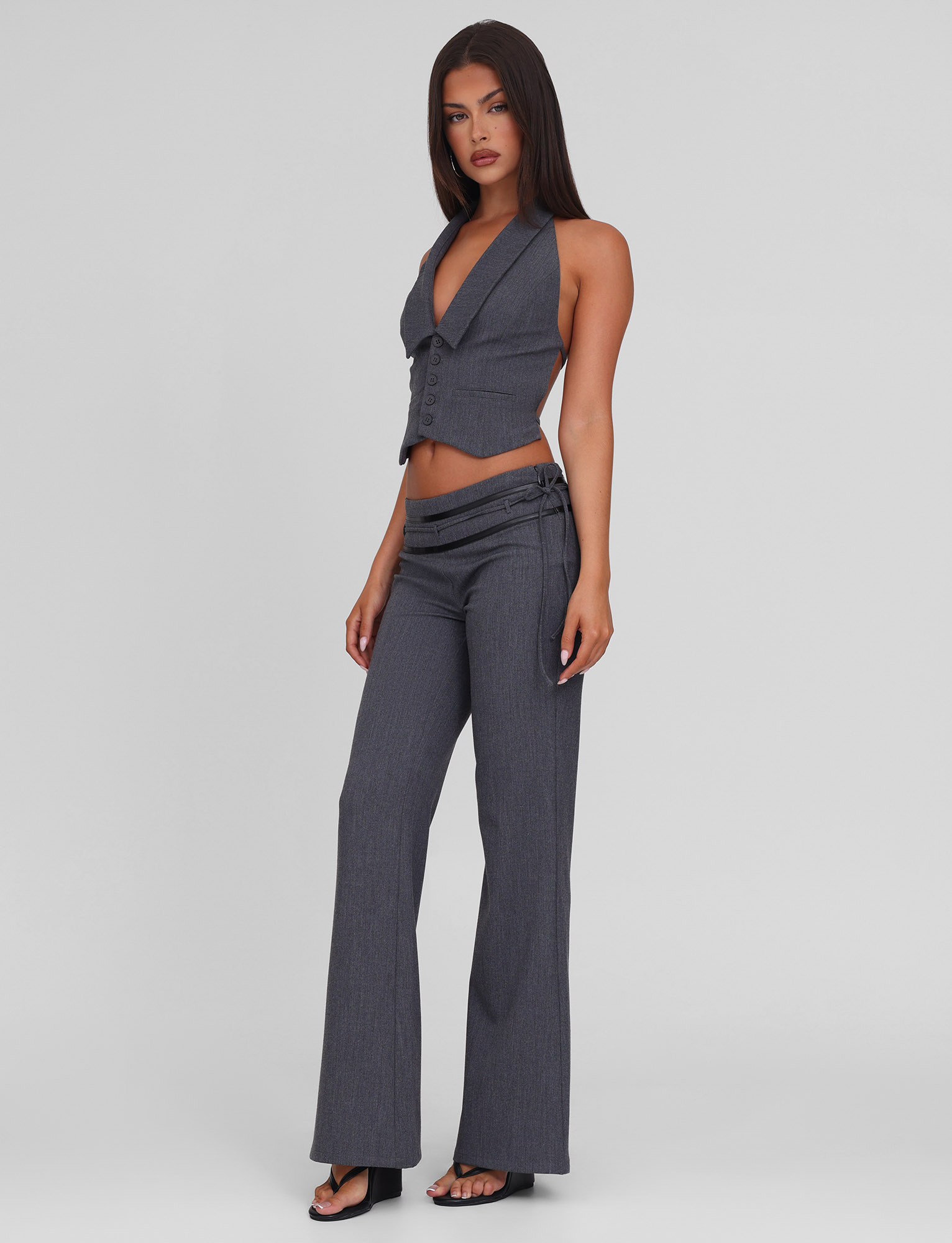 A woman stands against a plain background wearing TIGERMIST's FEDRA PANT in grey—a high-waisted, flared style with slits and belt detail—paired with a sleeveless, deep V-neck vest with PU leather trim, all in soft touch suiting fabric and black heeled sandals.