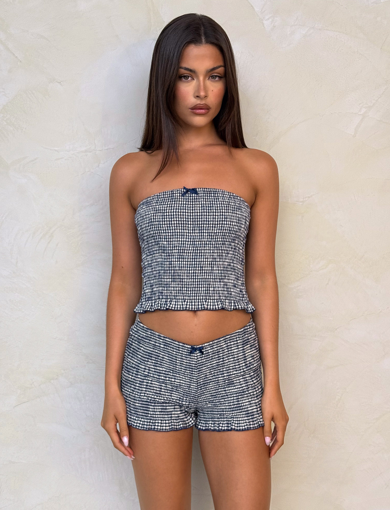 A woman with long brown hair wears TIGERMIST's ERYNN SHORTS in navy gingham, featuring ruffled leg openings and a smocked, strapless matching top, standing against a textured wall and looking directly at the camera with a neutral expression.