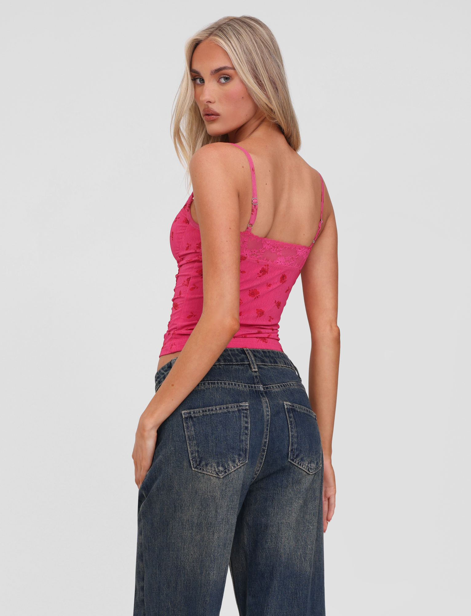 A woman with long blonde hair poses in the TIGERMIST KHALIA TOP - DITSY FLORAL, a pink floral camisole with adjustable straps, paired with loose dark blue jeans, against a plain white background.